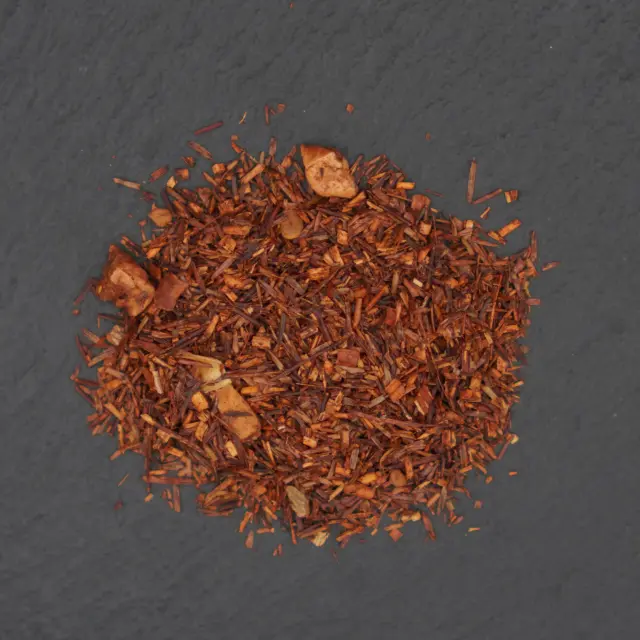 Un munte de ceai rooibos uscat, cu bucăți de măr copt și condimente, pe o suprafață închisă la culoare.