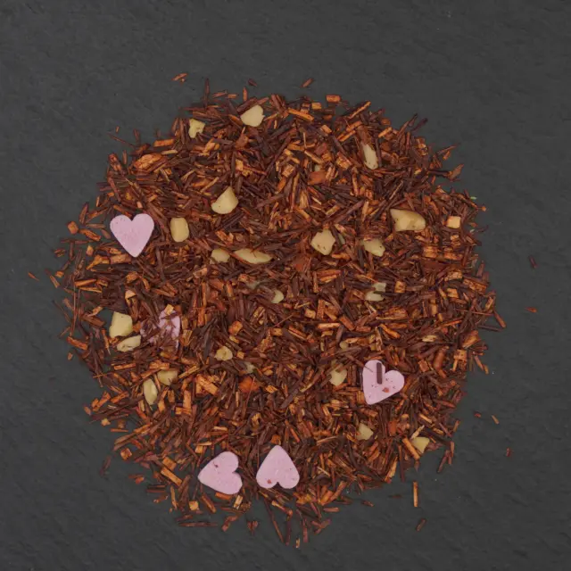 Un amestec de ceai rooibos cu bucăți de migdale și inimioare roz, așezat pe o suprafață închisă la culoare.