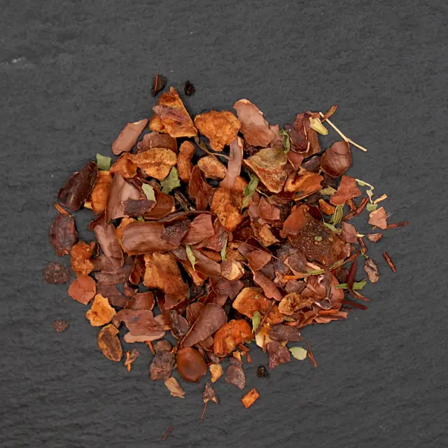 Un amestec de ceai Caramellino, cu bucăți de cacao, fructe uscate și frunze verzi, pe o suprafață închisă la culoare.