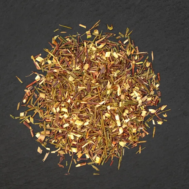 O grămadă de ceai rooibos verde, cu frunze tăiate fin, de culoare verde-maroniu, pe o suprafață închisă la culoare.